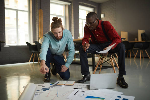 Full length portrait of two contemporary businessmen laying documents on floor while planning startup project in loft styled office, copy space
